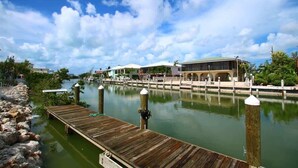 Una propiedad frente al agua con un muelle de madera que se adentra en aguas tranquilas, un edificio con techo verde y un cielo azul despejado con algunas nubes.