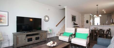 Ein Wohnzimmer mit Fernseher, Couchtisch aus Holz und zwei weiße Sessel mit grünen Kissen.
