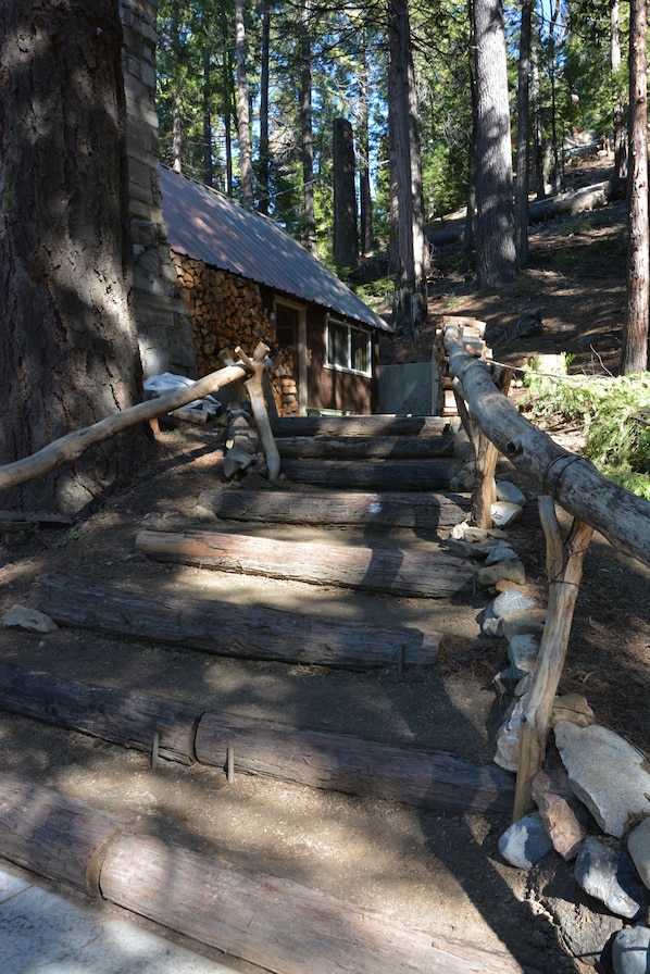 A rustic wooden staircase leading to a stone building with a metal roof, surrounded by trees.