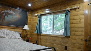 A wooden cabin room with a bed, a lamp, a window with curtains, and a painting on the wall.
