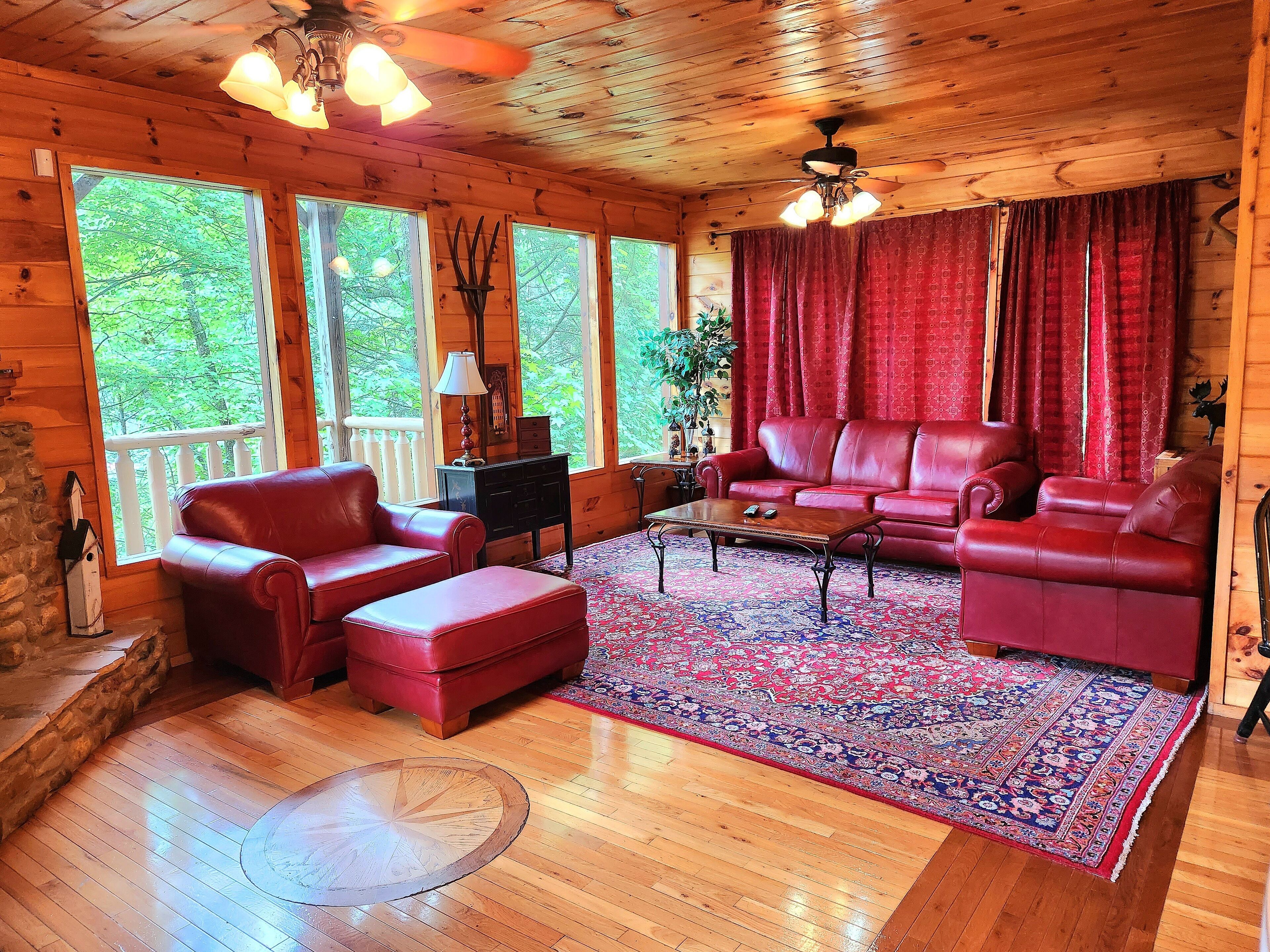 A cozy cabin interior with a wooden floor, a red leather sofa, a coffee table, and a large window with a view of greenery.