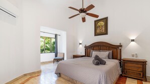 Ein Schlafzimmer mit einem Bett, einem Deckenventilator, einem Nachttisch, einem Sessel und einem Fenster mit Blick auf Grünflächen.