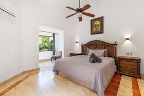 Une chambre à coucher comprenant un lit, un ventilateur de plafond, une table de chevet, une chaise et une fenêtre donnant sur un paysage verdoyant.