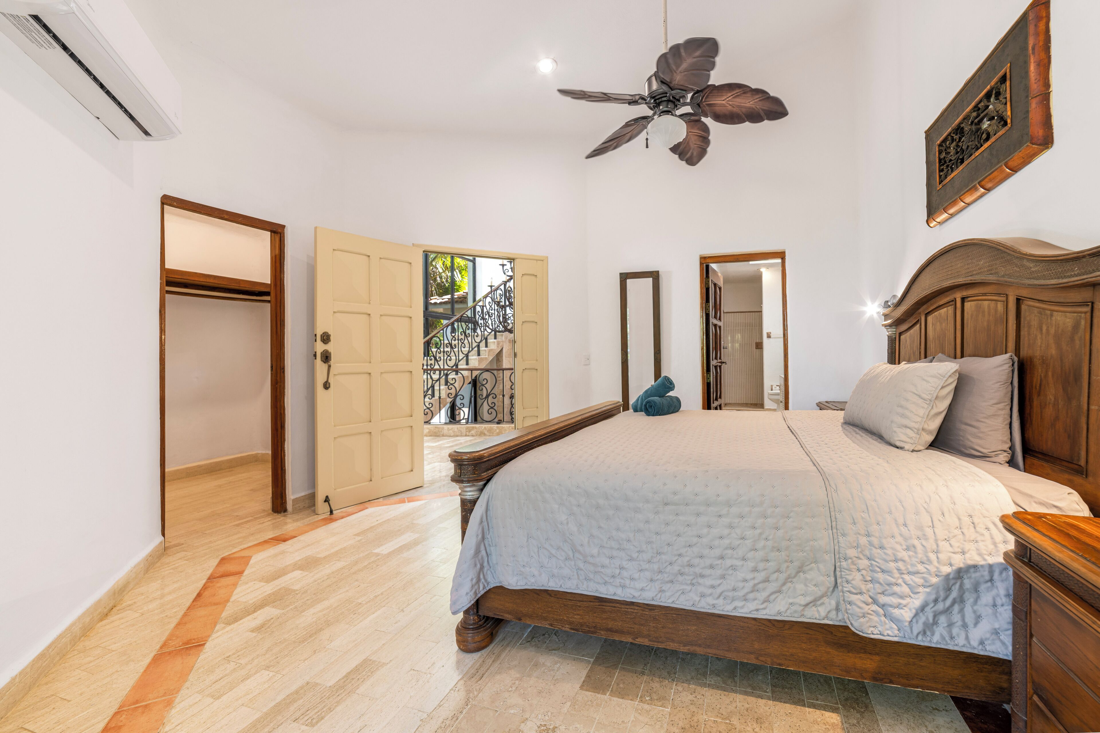 Une chambre à coucher avec un lit, une tête de lit en bois, un ventilateur de plafond et une vue sur un balcon.