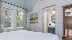 A bedroom with a large bed, a painting on the wall, a mirror, and a wooden bench with a pillow. - "Tupelo Honey" New Upscale Home With Magnificant Golf Course Views; screened in porch (Bald Head Island)