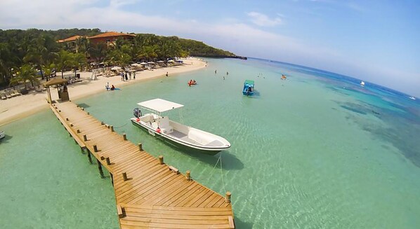 A wooden pier leading to a small boat docked at a beach with clear turquoise water and a few people in the distance. - ONLY Beachfront Suite @ Infinity Bay Resort - 24hr Service, Pools, Spa, Luxury! (West Bay Beach)
