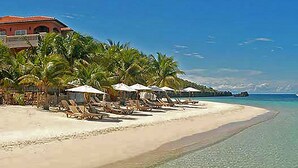 A beach with sun loungers and umbrellas, a building with a red roof, and a clear blue sky. - ONLY Beachfront Suite @ Infinity Bay Resort - 24hr Service, Pools, Spa, Luxury! (West Bay Beach)