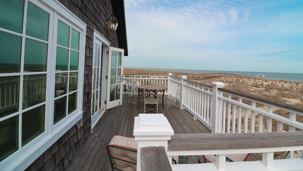 A wooden deck with a white railing, a table, and chairs, overlooking a beach with dunes. - Oceanfront Historic Beach Cottage | Steps from Beach (Tybee Island)