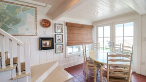 A dining area with a wooden table and chairs, a map on the wall, and a staircase with white railings. - Oceanfront Historic Beach Cottage | Steps from Beach (Tybee Island)