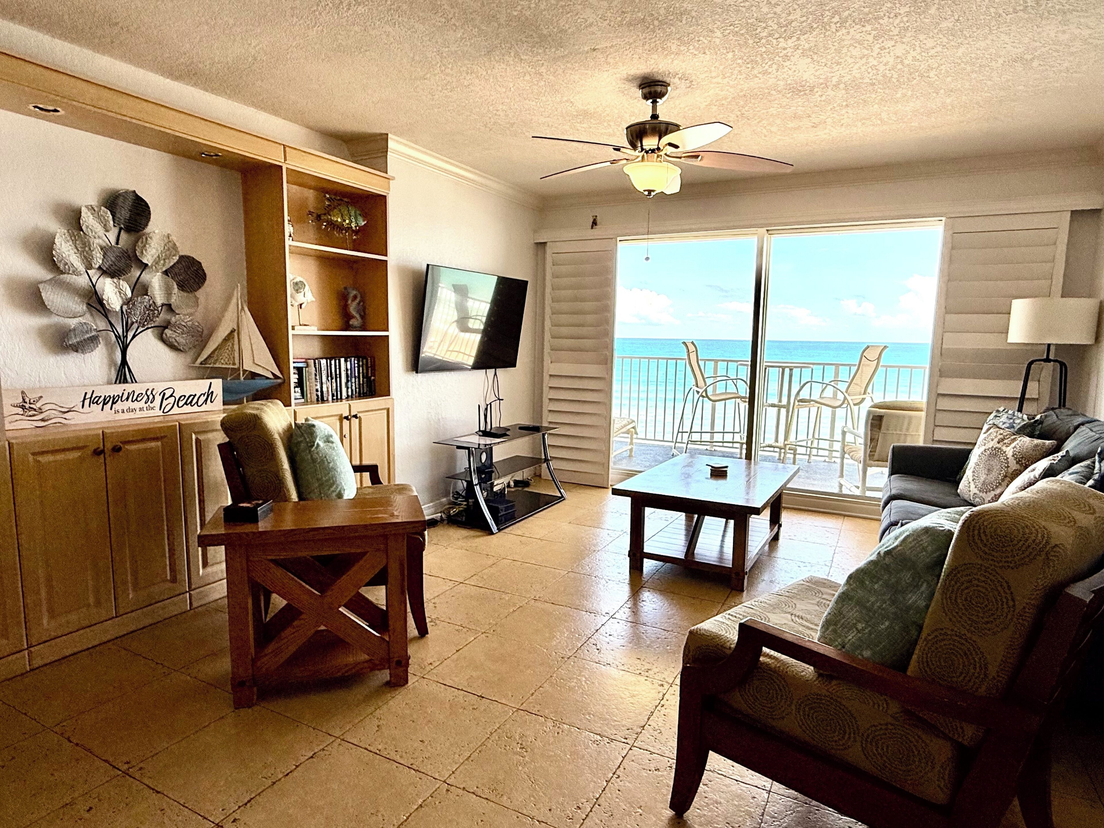 A living room with a TV, wooden furniture, a ceiling fan, and a view of the ocean.