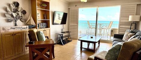 A living room with a TV, wooden furniture, a ceiling fan, and a view of the ocean.