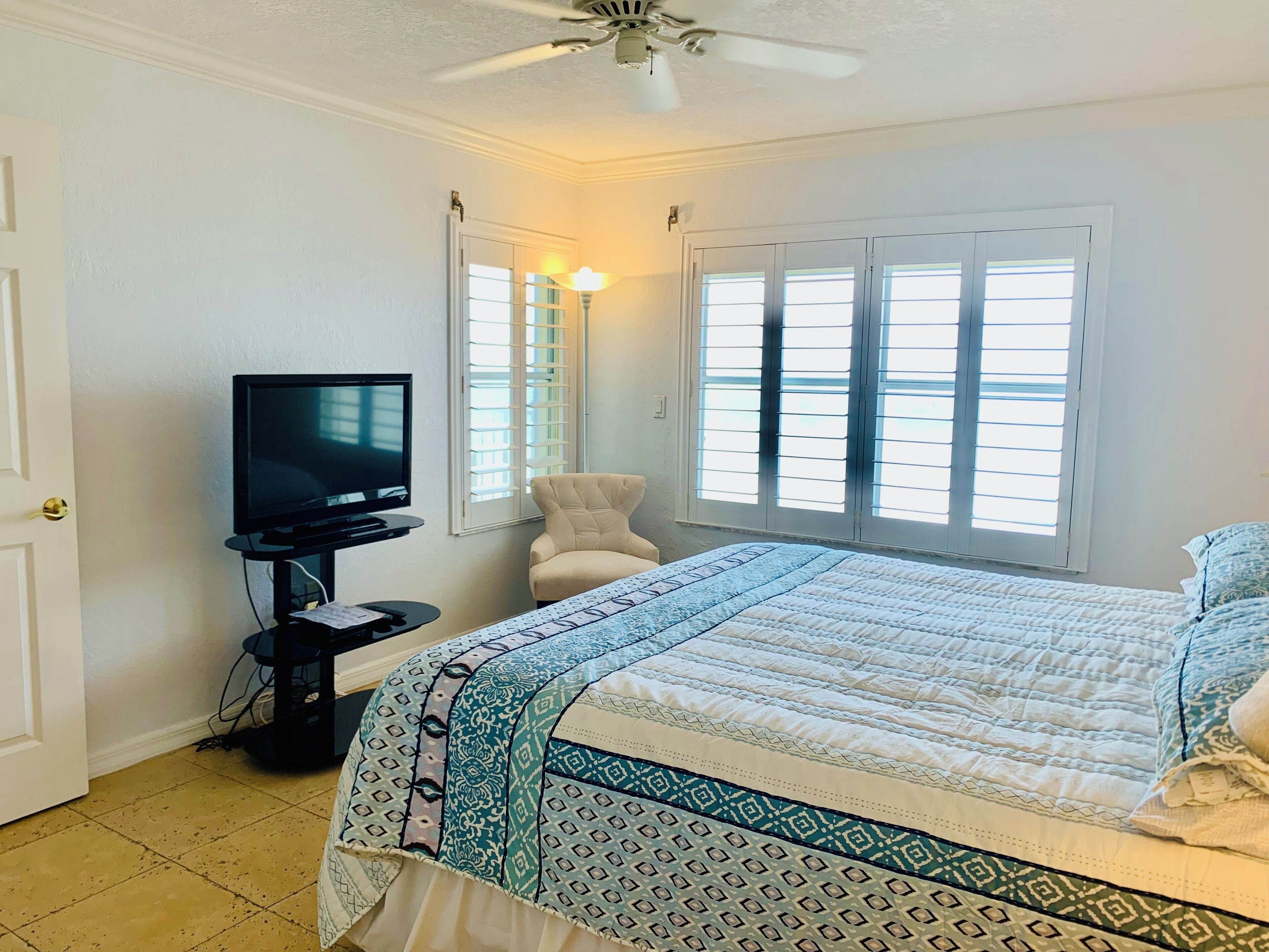 Un dormitorio con una cama, un televisor sobre un mueble, una silla y una ventana con persianas.