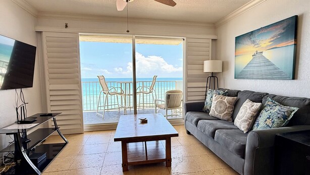 A living room with a TV, a coffee table, a sofa with cushions, a painting, and a view of the ocean through a sliding door. - Large Oceanfront 2BD/2BA, Sleeps 6, Pool, Kitchen, W/D, Parking, Ormond/Daytona (Ormond Beach)