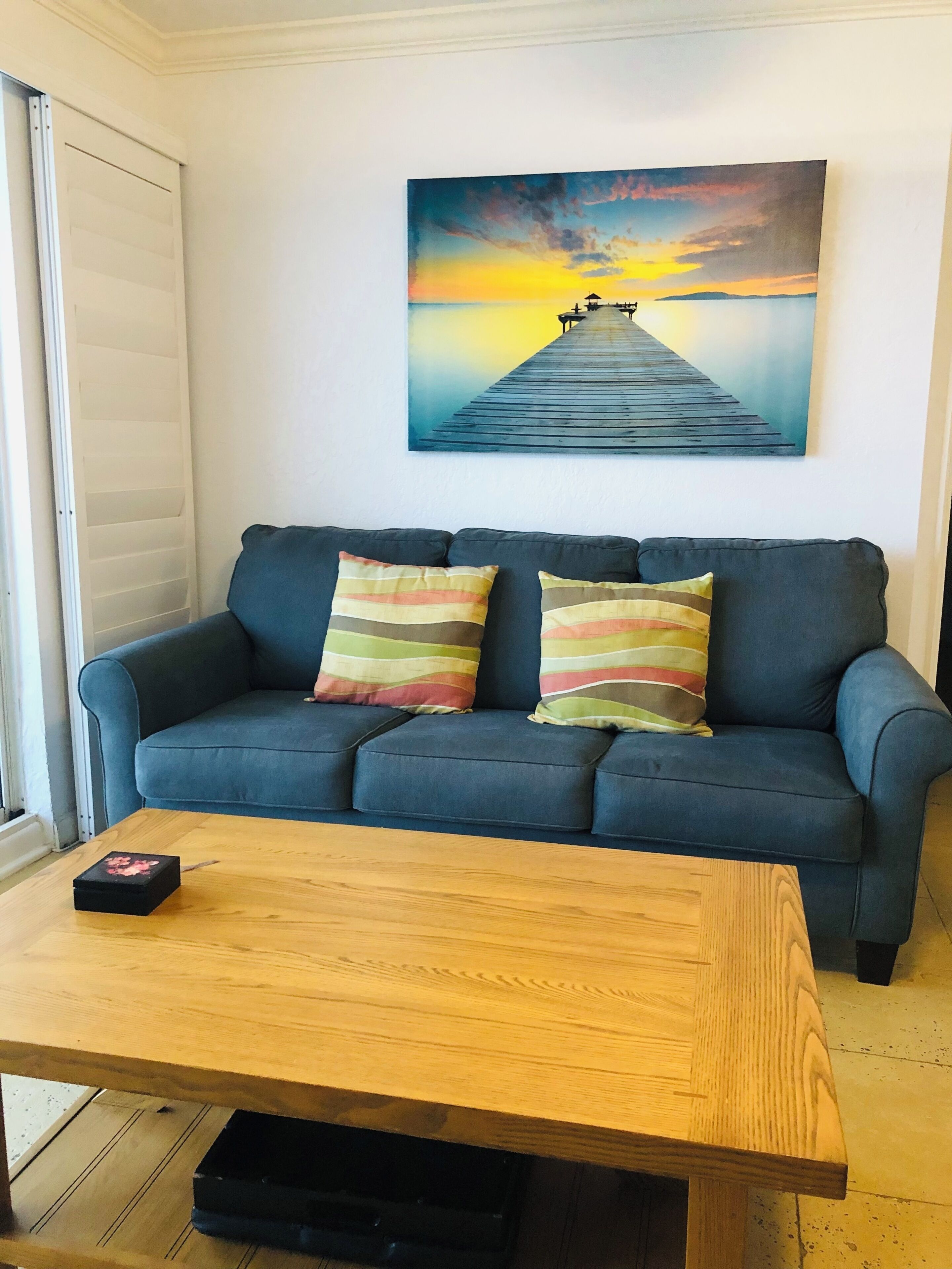 Una sala de estar con un sofá azul, dos almohadas a rayas, una mesa de centro de madera y un cuadro de un muelle en la pared.