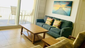A living room with a wooden table, a green sofa with striped pillows, a patterned armchair, and a painting of a pier on the wall. - Large Oceanfront 2BD/2BA, Sleeps 6, Pool, Kitchen, W/D, Parking, Ormond/Daytona (Ormond Beach)