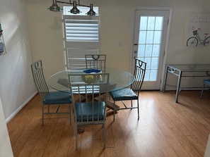 A dining area with a glass table, metal chairs, a window with blinds, and a door.