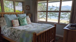 Un dormitorio con cama, cabecera de madera, ventana con vistas a una playa y montañas, y una mesita de noche con lámpara.