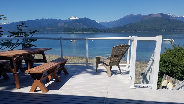 Terraza de madera con mesas y sillas de picnic que da a un lago con barcos y montañas al fondo.