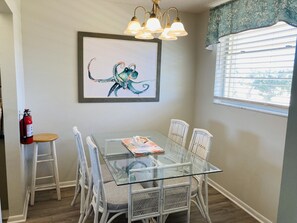 A dining area with a glass-top table, white wicker chairs, a framed octopus artwork, a fire extinguisher, and a window with blinds.