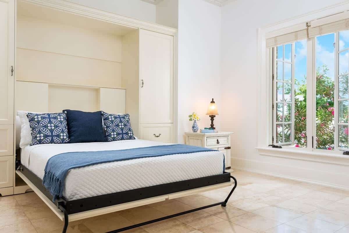 A bedroom with a bed, bedside table, lamp, and a window with a view of greenery.