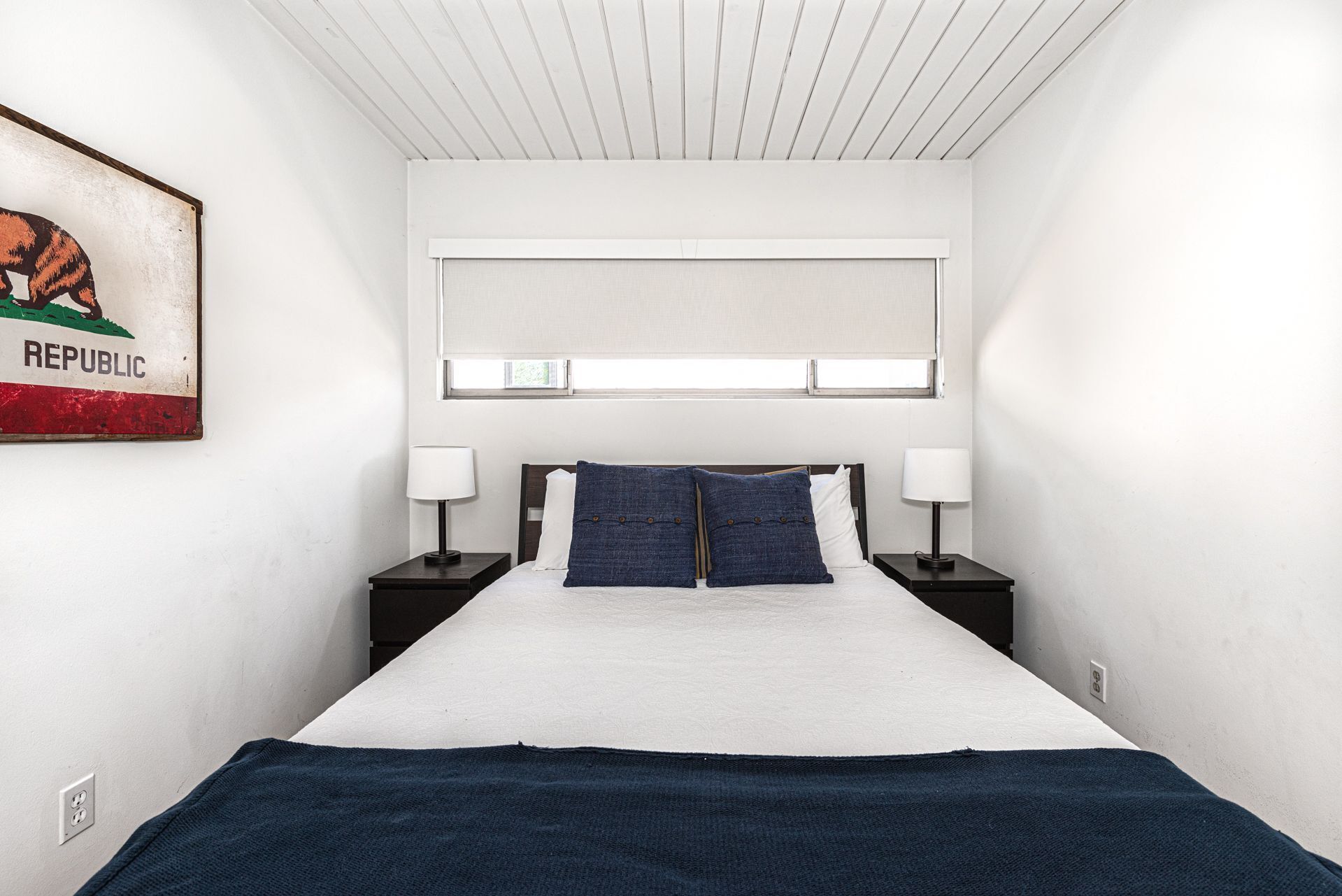 A bedroom with a bed, two bedside tables with lamps, a headboard, a window with blinds, and a wall-mounted painting.