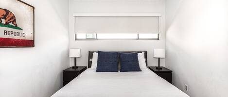 A bedroom with a bed, two bedside tables with lamps, a headboard, a window with blinds, and a wall-mounted painting.