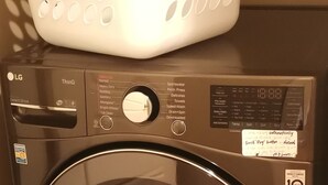 A front-loading LG washing machine with a control panel, a white laundry basket, and a shelf with cleaning supplies. - Self Entrance 2 BR, Whole Suite, Close To White Rock Beach & US Border, NEW (Surrey)