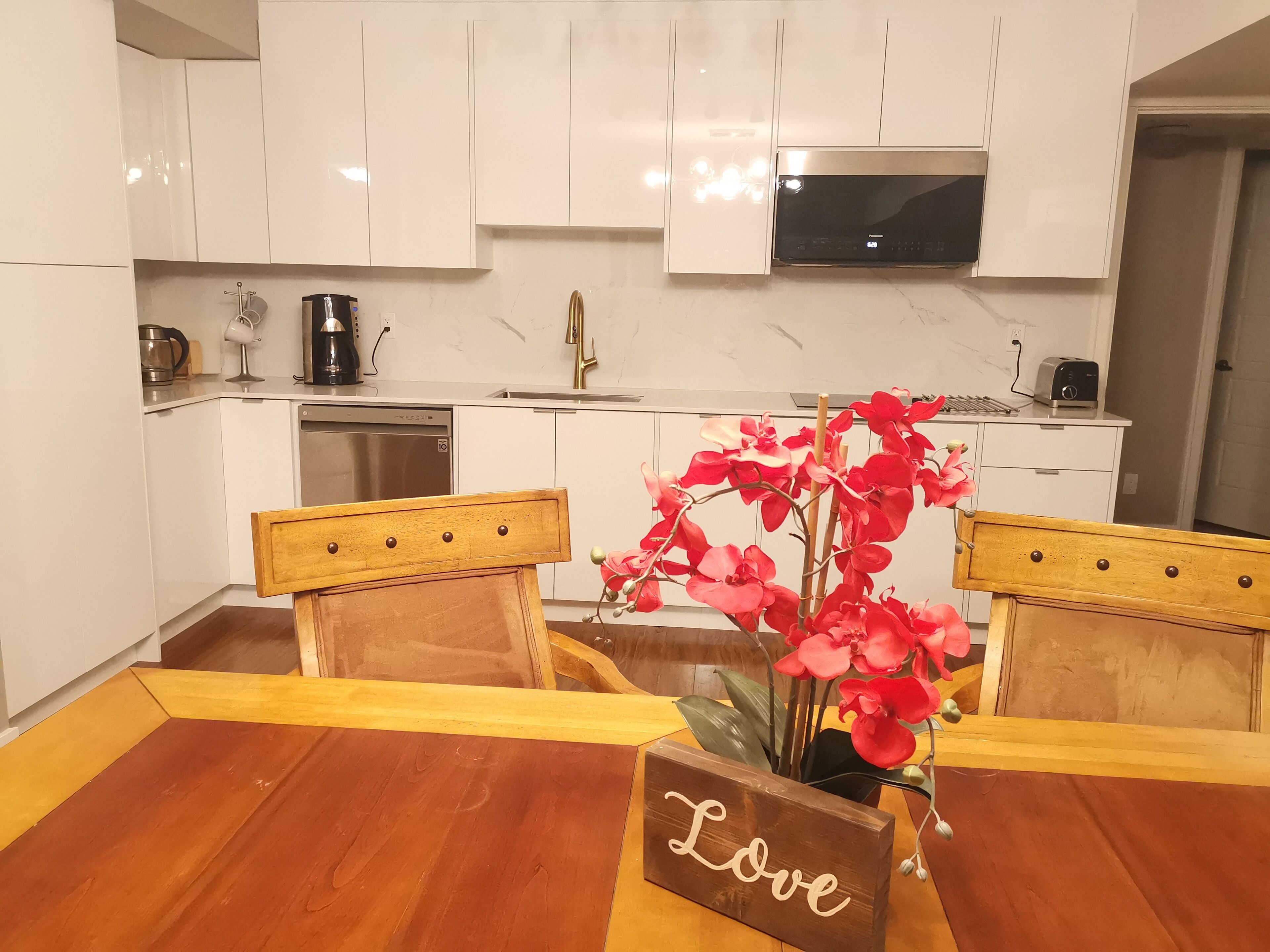 A modern kitchen with white cabinets, a built-in oven, and a microwave. There is a wooden dining table with a sign that reads "Love" and a vase with red flowers.