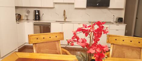 A modern kitchen with white cabinets, a built-in oven, and a microwave. There is a wooden dining table with a sign that reads "Love" and a vase with red flowers.