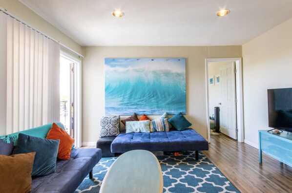 A living room with a large ocean wave painting, a sofa with colorful cushions, a coffee table, and a TV on a stand.