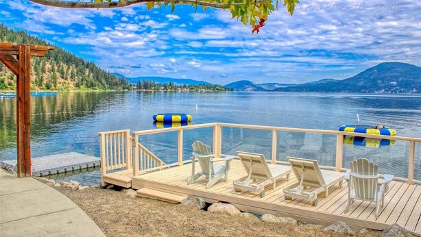 Une terrasse en bois, dotée de chaises longues et d’une rambarde en verre, offre une vue sur un lac, avec des montagnes en toile de fond.