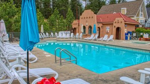 Une piscine avec des chaises longues blanches et un parasol bleu. On aperçoit, au fond, un bâtiment avec un toit rouge.