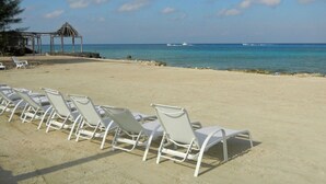 Une plage avec des chaises longues blanches alignées en rangées, une petite construction et des eaux bleues et calmes, avec des bateaux visibles au loin.