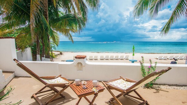 Une terrasse avec deux chaises en bois et une table, d’où l’on a une vue dégagée sur une plage, l’océan et les bateaux au loin.