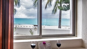 Une fenêtre donnant sur une plage et l’océan, un livre, deux verres de vin rouge, une bougie et quelques coquillages posés sur un rebord.