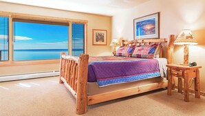 Ein hölzernes Bett mit einer farbenfrohen Steude, ein Nachttisch mit einer Lampe und ein großes Fenster mit Blick auf das Meer.