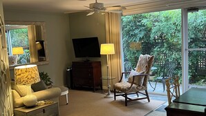 A living room with a television, a chair, a table, and a view of greenery outside.