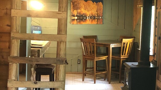 Interior rústico de una cabaña con una escalera de madera, una mesa de comedor con sillas, un cuadro en la pared y un estufa de leña.
