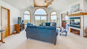 A spacious living room with a large gray sofa, a television, and a ceiling fan. There are large windows providing a view of the outdoors.