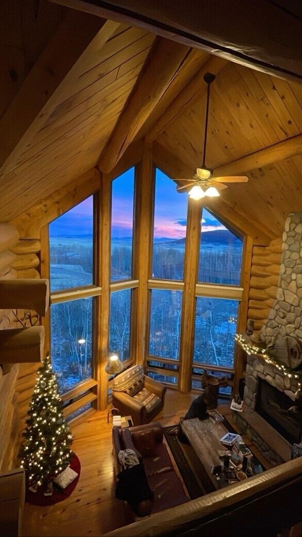 A cozy cabin interior with a large window, a fireplace, a Christmas tree, and a ceiling fan.