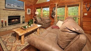 Ein gemütliches Blockhaus-Interieur mit Kamin, Ledersofa und großen Fenstern mit Blick auf einen Wald.