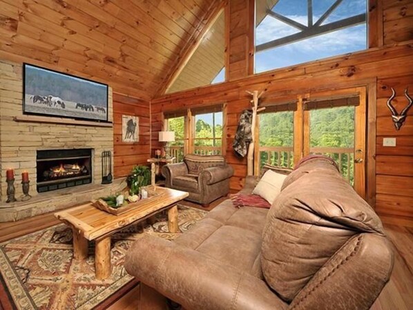 A cozy log cabin interior with a fireplace, leather sofa, and large windows overlooking a forest.