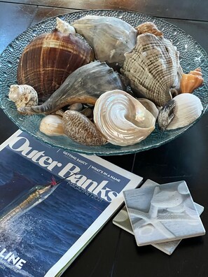 A glass bowl filled with various seashells on a table next to a magazine titled "Outer Banks Magazine 2021".