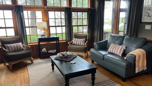 Een gezellige woonkamer met een donkere leren bank, twee fauteuils, een houten salontafel en een groot raam met gordijnen.