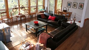 Een ruime woonkamer met een zwarte leren bank, houten stoelen, een salontafel en een eettafel. Grote ramen bieden uitzicht op de omgeving.