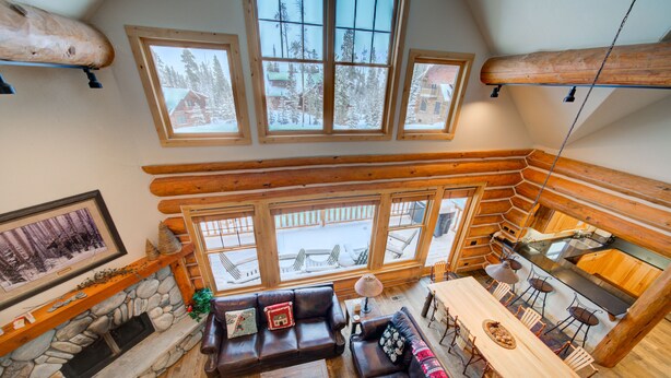 A cozy log cabin interior with a stone fireplace, wooden furniture, and large windows offering a view of the snowy outdoors.