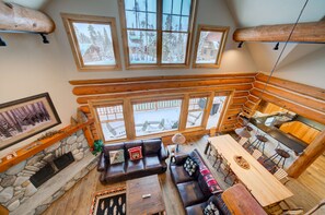 L’intérieur chaleureux d’un chalet en rondins, avec une cheminée en pierre, des meubles en bois et de grandes fenêtres donnant sur le paysage enneigé.