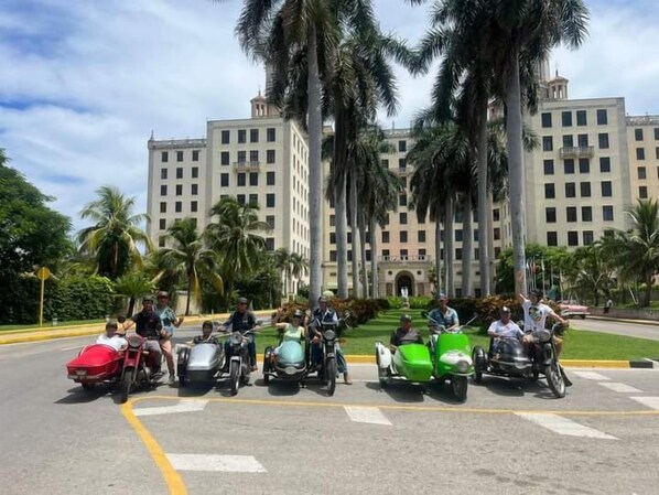 Motorcyclists gather in front of a grand building surrounded by lush greenery.