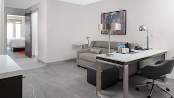 A modern hotel room with a grey sofa, a desk with a chair, a standing lamp, and a painting on the wall.