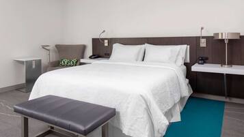 A modern hotel room with a large bed, a bench, a bedside table, a lamp, and a small bedside table with a phone and a charger.